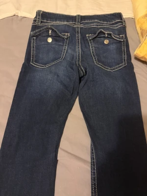 Mörkblå jeans med kontrastsömmar - Snygga mörkblå jeans med raka ben och vita kontrastsömmar. Jeansen har två bakfickor med lock och silverfärgade knappar. Klassisk femficksmodell i mjukt denimtyg som passar perfekt till en avslappnad stil.