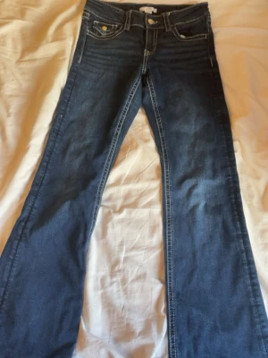 Mörkblå bootcut jeans med kontrastsömmar - Snygga mörkblå jeans med bootcut-modell och vita kontrastsömmar. Klassisk femficksdesign och detaljerade bakfickor med knappar. Jeansen har normal midja och är tillverkade i ett mjukt denimtyg som ger en bekväm passform.