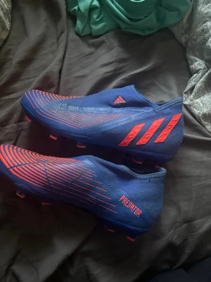 Blåa Adidas Predator fotbollsskor - Säljer ett par blåa Adidas Predator fotbollsskor med röda detaljer och mönster. Skorna har en strumpliknande passform utan snörning och är designade för spel på gräs. Tillverkade i syntetmaterial för bra grepp och kontroll på planen.