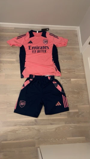 Arsenal träningsset rosa/marinblå Adidas - Säljer ett Arsenal träningsset från Adidas med t-shirt och shorts. Tröjan är rosa med marinblå detaljer och tryck, medan shortsen är marinblå med rosa detaljer och Arsenal-logga. Setet är gjort i lätt och snabbtorkande material, perfekt för fotbollsträning.