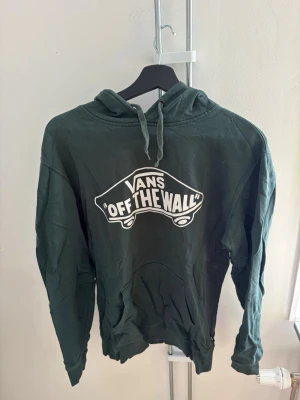 Grön hoodie från Vans med tryck - Mörkgrön hoodie från Vans med stort vitt 'Off The Wall'-tryck på bröstet. Klassisk modell med huva och dragsko samt känguruficka framtill. Perfekt för en avslappnad streetwear-look.