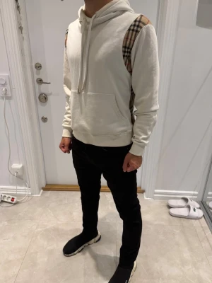 Vit Burberry hoodie med rutiga detaljer - Säljer en vit Burberry hoodie med huva och klassiska beige, svarta och röda rutiga paneler på axlar och sidor. Hoodien har känguruficka framtill och ribbade muddar vid ärmslut och nederkant. Perfekt för dig som gillar stilrena plagg med en twist.