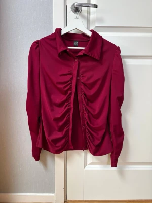 Vinröd blus med rynk framtill - Stilren vinröd blus med långa ärmar och markerad krage. Blusen har knappar framtill och snygga rynkdetaljer längs knappraden som ger en trendig look. Tillverkad i ett mjukt, stretchigt material som sitter bekvämt.