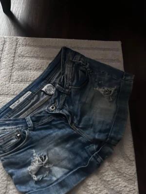 Vintage jeans shorts  - Säljer ett par blå jeansshorts med slitna detaljer och råa kanter. Shortsen har klassisk femficksdesign, knappgylf och en cool, avslappnad look. Perfekta för varma dagar och ger en edgy vibe till din outfit.