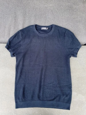 Mörkblå stickad t-shirt - Mörkblå stickad t-shirt från Dressmann med korta ärmar och rund hals. Tröjan har en diskret strukturstickning och ribbade muddar vid ärmslut och nederkant. Perfekt för dig som vill ha en enkel men stilren look.