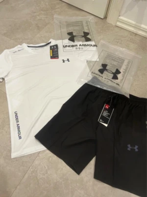 Under Armour träningsset svart/vit - Snyggt träningsset från Under Armour med en vit t-shirt och svarta shorts. T-shirten har tryckt logga på bröstet och text längs sidan, tillverkad i lätt och ventilerande material med HeatGear-teknologi. Shortsen har elastisk midja och logga på benet.