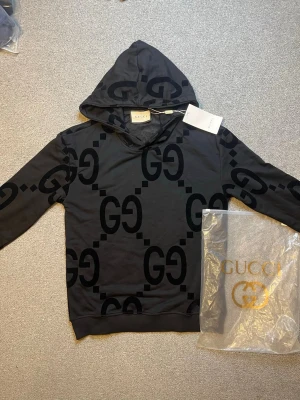 Svart hoodie med GG-mönster från Gucci - Snygg svart hoodie från Gucci med stort GG-mönster över hela tröjan. Hoodien har huva och ribbade muddar vid ärmslut och nederkant. Perfekt för dig som gillar exklusiva streetwear-plagg och vill sticka ut med en ikonisk design.