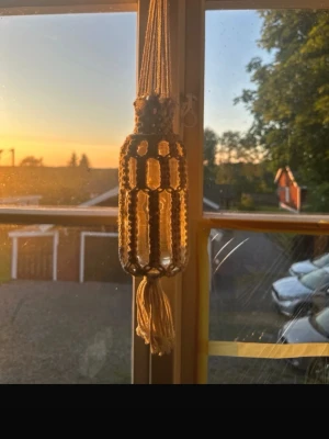 Makramé ampel i beige bomull - Handknuten makramé ampel i beige bomullssnöre med dekorativt mönster och lång tofs nedtill. Hängs upp med en trä-ring och passar perfekt för att ge boho-vibbar till rummet. Snygg detalj att ha i fönstret eller på väggen.