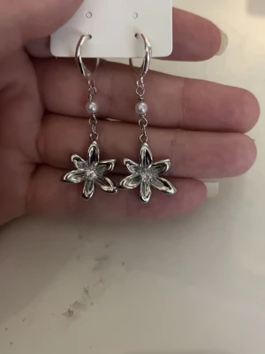 Silvriga blommiga örhängen med pärlor - Säljer ett par silvriga örhängen med hängande blommor i metall och små vita pärlor ovanför blomman. Örhängena har en elegant och modern design med blommotiv och passar perfekt för dig som gillar detaljerade accessoarer.