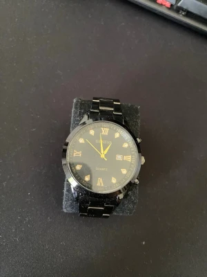 Svart klocka med gulddetaljer Geneva - Stilren svart klocka från Geneva med guldfärgade visare och romerska siffror på urtavlan. Länken är i blank svart metall och klockan har en klassisk rund form. Perfekt accessoar för dig som gillar en elegant look.
