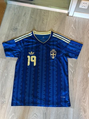 Blå Sverige fotbollströja - Säljer en blå fotbollströja från Adidas med Sveriges landslagsemblem och nummer 19 i gult. Tröjan har korta ärmar, gul krage och tre gula ränder på axlarna. Namnet Eliasson står på ryggen och materialet är lätt och andas.