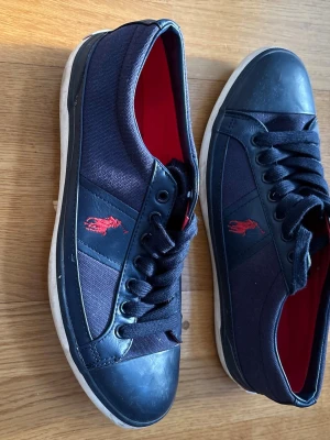 Svarta Polo Ralph Lauren sneakers - Snygga blåa sneakers från Polo Ralph Lauren med röd logga på sidan och röd insida. Skorna har lågt skaft, svarta snören och detaljer i både textil och läderimitation. Perfekta för en stilren och avslappnad look. Pris går att diskuteras vid snabb affär.