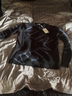 Svart stickad tröja från Valentino - Snygg svart stickad tröja från Valentino med stora blå VLTN-bokstäver över hela tröjan. Tröjan har rund halsringning och långa ärmar. Perfekt för dig som gillar exklusiva och trendiga plagg med tydlig logga.