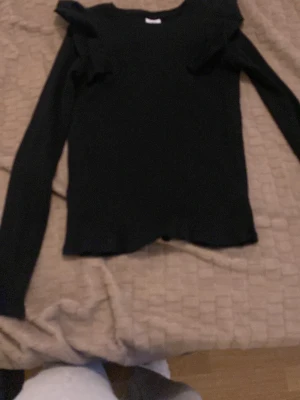 Svart långärmad topp från Zara - Snygg svart långärmad topp från Zara med volangdetaljer vid axlarna. Toppen är ribbad och har en kroppsnära passform som ger en stilren look. Perfekt att matcha med jeans eller kjol för en trendig outfit.