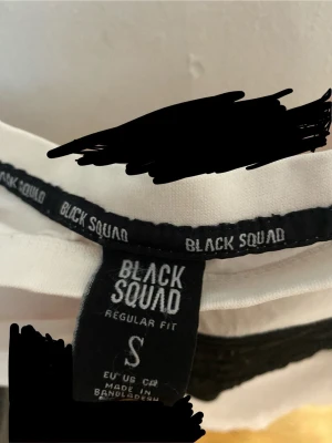 Vit t-shirt från Black Squad - Snygg vit t-shirt från Black Squad med regular fit och ett stort svart tryck på bröstet. Klassisk rund hals och korta ärmar. Perfekt till jeans eller shorts för en clean och modern look.