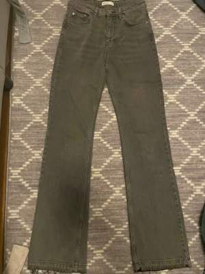 Grå raka jeans med rå kant - Säljer ett par grå jeans med rak passform och hög midja. Jeansen har fem fickor, bälteshällor och rå kant längst ner vid bensluten för en trendig look. Perfekta till sneakers eller boots.