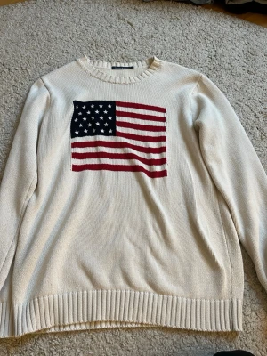 Vit stickad tröja Brandy Melville - Vit stickad tröja från Brandy Melville med amerikansk flagga på bröstet. Tröjan har rund halsringning, ribbade muddar och lång ärm. Perfekt för dig som gillar klassisk och avslappnad stil.