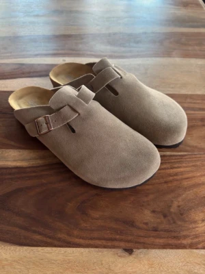 Birkenstock Boston taupe - Säljer ett par snygga beige tofflor från Birkenstock i mjuk mocka. De har en klassisk slip-in design med justerbart spänne i metall och svart sula. Perfekta för dig som vill ha både stil och komfort hemma eller ute.