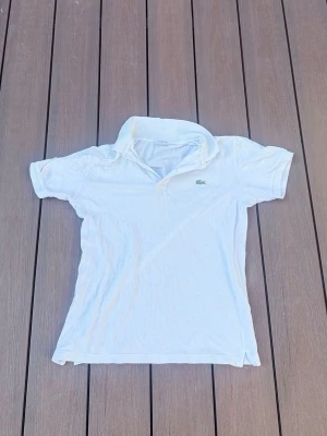 Vit pikétröja från Lacoste - Klassisk vit pikétröja från Lacoste i slim fit-modell. Tröjan har korta ärmar, krage och knäppning framtill samt den ikoniska gröna krokodilloggan på bröstet. Perfekt till en clean och sportig stil.