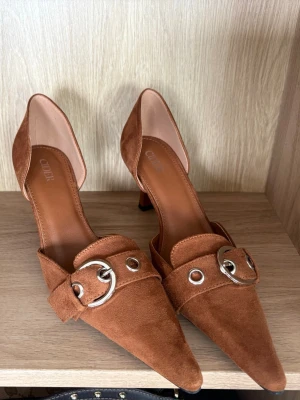 Bruna mockapumps med metallspänne - Snygga bruna pumps i mocka från Cider med spetsig tå och låg klack. Oanvända säljer då jag var osäker på storlek och de är förstora för mig som har 37,5. Men super super snygga 