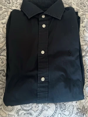 Svart skjorta från Polo Ralph Lauren - Klassisk svart skjorta från Polo Ralph Lauren med knappar framtill och diskret broderad logga på bröstet. Skjortan har en stilren krage och är tillverkad i bomull, perfekt för en clean och tidlös look.