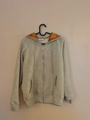 Grå zip hoodie från Burberry - Snygg grå zip hoodie från Burberry med klassiskt rutigt foder i huvan. Tröjan har dragsko i huvan, två fickor fram och ribbade muddar vid ärmar och nederkant. Perfekt för en avslappnad och trendig look.
