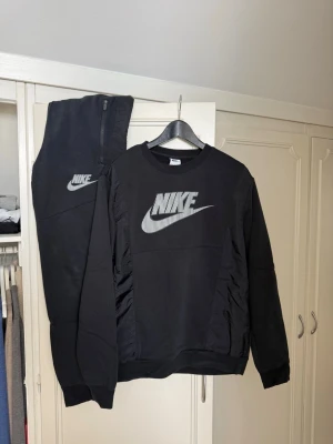Svart Nike loungewear set M - Snyggt svart loungewear set från Nike med matchande sweatshirt och byxor. Båda plaggen har Nike-logga i vitt framtill. Tröjan har rund hals och muddar vid ärmslut och midja, byxorna har resår i midjan och dragkedja upptill. Perfekt för chill dagar hemma.