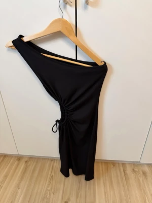 Svart oneshoulder klänning med snörning - Trendig svart oneshoulder klänning med snörning i sidan som ger en snygg rynkeffekt. Kort modell med tight passform, perfekt för dig som vill sticka ut. Tillverkad i ett mjukt och stretchigt material.