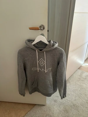 Grå hoodie från Gant med logga - Snygg grå hoodie från Gant med vit logga på bröstet och vita dragsnören i huvan. Klassisk känguruficka framtill och mjukt material!!🐘🐘