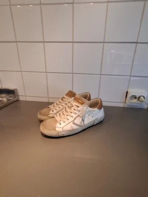 Philippe model skor - Säljer ett par vita phillip model med bruna detaljer på häl och plös. Skorna har klassisk låg profil, vita snören och sula samt diskreta sömmar. Materialet är läder eller konstläder med mockaliknande partier på tån. Perfekta för en avslappnad streetstil.
