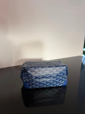 Blå mönstrad necessär med dragkedja Goy  - Snygg blå necessär med vitt grafiskt mönster och silverfärgad dragkedja. Väskan har detaljer i blått konstläder och praktisk rektangulär form. Perfekt för att organisera småsaker eller smink i väskan.