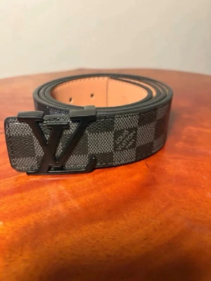 Louis Vuitton rutigt svart bälte - Snyggt svart bälte från Louis Vuitton med klassiskt rutigt mönster och stor LV-logga i metall som spänne. Bältet har en exklusiv känsla och är gjort i skinn med detaljer i mörkgrått och svart. Perfekt accessoar för att lyfta din outfit.