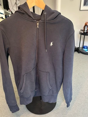 Mörkblå zip hoodie från Polo Ralph Lauren - Mörkblå hoodie med dragkedja från Polo Ralph Lauren. Klassisk design med huva, dragkedja och två fickor framtill. Ikoniska vita Polo-loggan broderad på bröstet. Perfekt för en avslappnad och stilren look.
