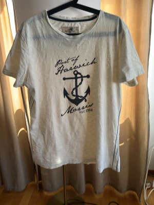 Vit Morris t-shirt med marinblått ankare - Vit t-shirt från Morris med marinblått tryck av ankare och texten 'Port of Harwich'. Klassisk rund halsringning och korta ärmar. Tillverkad i bomull, perfekt för en avslappnad och stilren look.