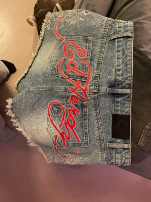 Ed Hardy jeansshorts med broderi - Säljer ett par ljusblå jeansshorts från Ed Hardy med råa kanter och stort rött broderi på baksidan. Klassisk femficksmodell med bälteshällor och knappgylf. Snygg slitning framtill och cool detalj med broderat mönster på ena benet.