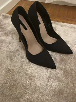 Svarta klassiska pumps med spetsig tå - Säljer ett par svarta pumps med spetsig tå och hög smal klack. Skorna har en stilren design och är tillverkade i ett tygmaterial med matt finish. Insidan är ljus och sulan har en neutral ton. Perfekta för dig som gillar en elegant look.