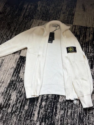 Vit stickad ziptröja Stone Island - Säljer en fräsch vit stickad ziptröja från Stone Island med ikonisk patch på ärmen. Tröjan har hög krage, dragkedja framtill och ribbade muddar. Perfekt för dig som gillar stilrena och exklusiva plagg. Storlek kan funka på S och M