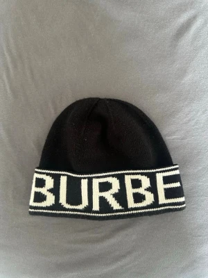 Svart mössa med vit Burberry-text - Snygg svart mössa med uppvikt kant och stor vit Burberry-logga runt hela kanten. Klassisk och stilren design som passar perfekt till streetwear eller för att lyfta din outfit. Stickad i mjukt material för skön känsla och bra passform.