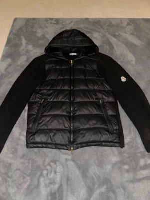 Moncler cardigan  - Snygg svart pufferjacka med huva och dragkedja framtill. Jackan har stickade svarta ärmar och två dragkedjeförsedda fickor på sidorna. Klassisk quiltad design på kroppen och en diskret logotyp på ena ärmen. Perfekt för kalla dagar.