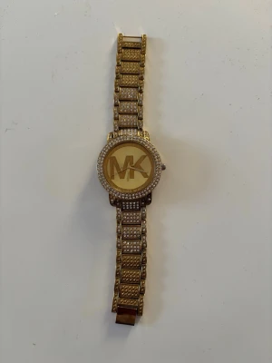 Guldig Michael Kors klocka med stenar - Snygg guldig klocka från Michael Kors med stora MK-bokstäver på urtavlan. Armbandet och boetten är dekorerade med glittrande stenar som ger en lyxig känsla. Perfekt accessoar för att lyfta din outfit. Behöver nytt batter