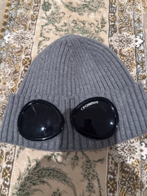 Grå goggle-mössa från C.P. Company - Säljer en grå ribbstickad mössa från C.P. Company med de ikoniska svarta goggle-linserna framtill. Mössan är tillverkad i mjukt material och har en uppvikt kant. Perfekt för dig som vill sticka ut med streetwear-stil.