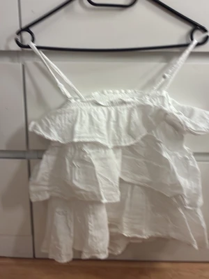 Vit volangig linnetopp - Söt vit linnetopp med tunna axelband och flera lager volanger. Toppen har en luftig och lätt känsla, perfekt för varma dagar. Den är ärmlös och har en avslappnad passform med romantiska detaljer.