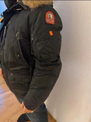 Svart Parajumpers parkas med päls - Svart parkajacka från Parajumpers med ikonisk gul rem vid kragen och patch på ärmen. Jackan har flera fickor, dragkedja och knappar samt en stor huva med päls. Perfekt för kalla dagar och riktigt snygg streetstil.Bra skick. Kvitto finns ☺️passar även för M, mörkblå 