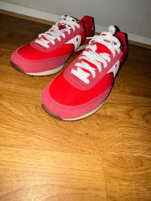 Röda Saucony sneakers med vita detaljer - Snygga röda sneakers från Saucony med vita detaljer och vita skosnören. Skorna har en klassisk låg siluett, beige sula och Saucony-logga på hälen. Ovandelen är i textil och mocka för en sportig look.