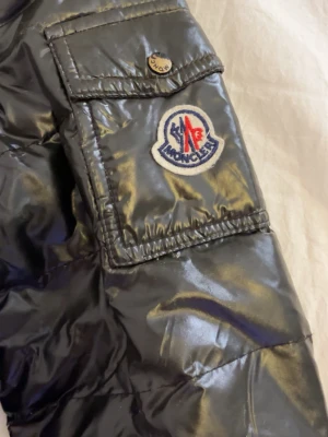 Svart pufferjacka från Moncler - Säljer en svart pufferjacka från Moncler med glansigt material , det är en väldigt gammal mansmodell är jag rätt så säker på men är inte helt hundra på att den är äkta då ja fick den av en bekant och man kan se att den är gammal därmed priset! Lite sliten uppe vid nacken och allmänt väl använd i skicket! Skulle säga att storleken är mellan xs och s.