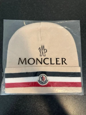 Beige Moncler mössa med logga - Snygg beige mössa från Moncler med broderad logga framtill och klassiska Moncler-ränder i svart, vitt och rött på uppviket. Tillverkad i mjukt material som håller dig varm och stilren under kalla dagar.Följ tt marknads.closet