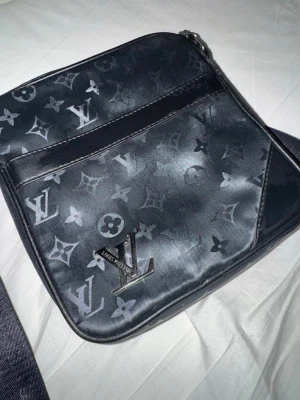Svart axelväska Louis Vuitton monogram - Snygg svart axelväska från Louis Vuitton med klassiskt monogrammönster och silverfärgad LV-logga framtill. Väskan har justerbar axelrem och är tillverkad i ett glansigt konstlädermaterial. Perfekt för att bära dina viktigaste grejer med stil.