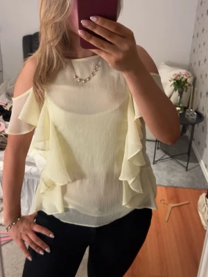 Ljusgul volangblus med cold shoulder - Söt ljusgul blus med volangdetaljer på sidorna och öppna axlar. Blusen är ärmlös och har en luftig, lätt transparent känsla. Perfekt för dig som gillar feminina detaljer och en somrig look.