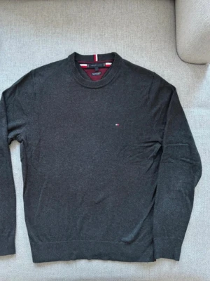 Mörkgrå tröja Tommy Hilfiger - Mörkgrå rundhalsad tröja från Tommy Hilfiger i en mjuk bomullsblandning med cashmere. Liten broderad logga på bröstet och ribbade muddar vid hals, ärmslut och nederkant. Perfekt för en stilren och avslappnad look.