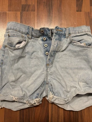 Ljusblå jeansshorts - Säljer ett par ljusblå jeansshorts från lager157 med hög midja och råa, fransiga benslut. Shortsen har klassisk femficksdesign och knappar i gylfen. Perfekta för sommaren och ger en avslappnad vibe.